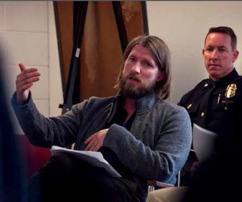 Brad Ray sits in a classroom with a police man behind him.