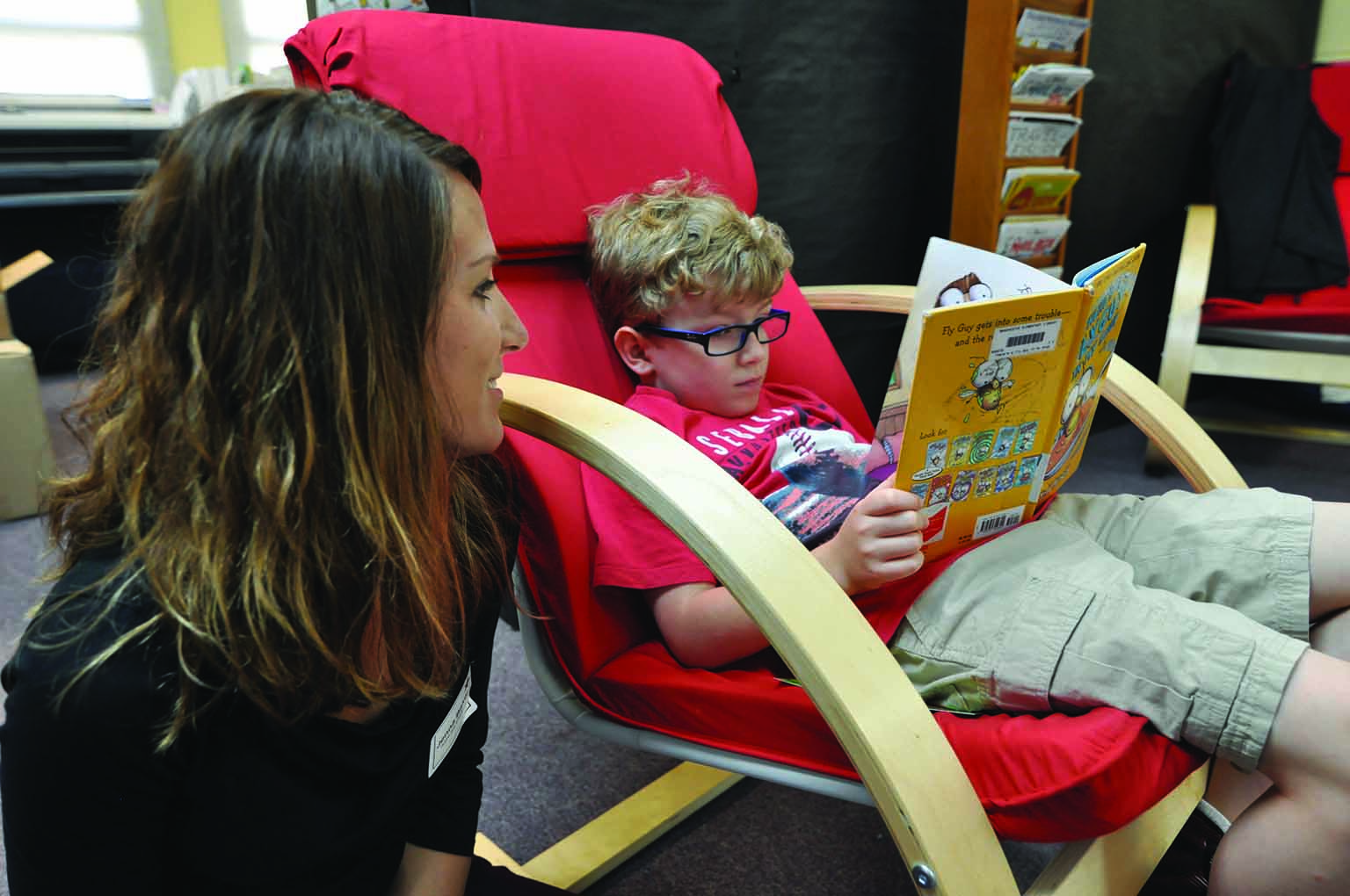 A little boy sits in a red chair and reads a picture book to a woman